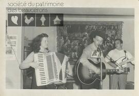 Dans un intérieur, devant une tapisserie, un groupe de musique avec deux guitares et un accordéon joue